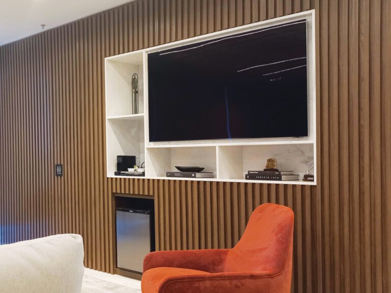 Parede de painel ripado executado pela Marcenaria Ensaios e Modelos, absorvendo fiação de Wi-Fi, TV, geladeira e ar-condicionado, com nichos acomodando os equipamentos.